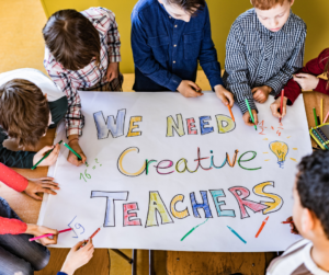 Children with Sign that Reads "We Need Creative Teachers"